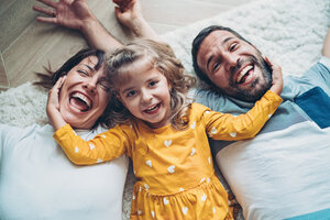 Eltern liegen mit ihrer Tochter auf dem Boden/Teppich und die Tochter umklammert dabei die Gesichter der Eltern mit der Hand | © pixelfit - Getty Images/iStockphoto